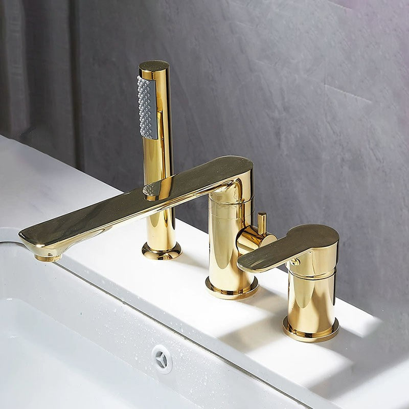 A modern three-hole bathroom faucet set with a polished gold finish, featuring a central spout and two separate handles, mounted on a white sink against a gray tiled wall.