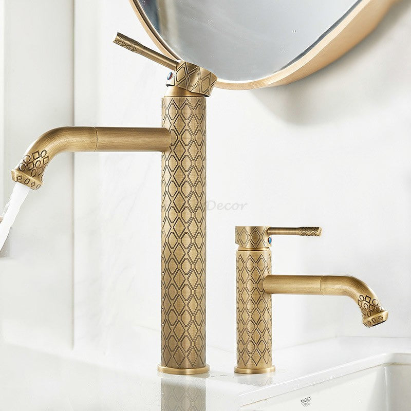 Two ornate brass bathroom faucets with a textured geometric pattern, shown installed on a white sink with water running from one faucet. A round mirror is partially visible in the background.