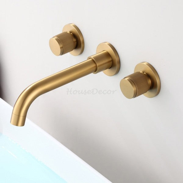 A three-hole wall-mounted bathroom faucet set in a brushed gold finish, featuring a curved spout and two round knobs, mounted on a white wall above a bathtub edge.