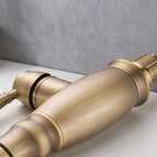 A close-up of a vintage-style faucet handle with a brushed antique brass finish, resting on a light surface against a textured gray background.