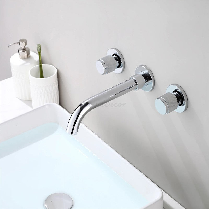 A modern wall-mounted three-hole sink faucet set with a polished chrome curved spout and two round control knobs, installed on a light gray bathroom wall above a white sink.
